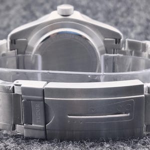 May include: A silver-toned wristwatch with a metal band and clasp. The watch is viewed from the back, showcasing the band's design and the clasp with the brand name "TUDOR" engraved. The watch is on a gray surface.