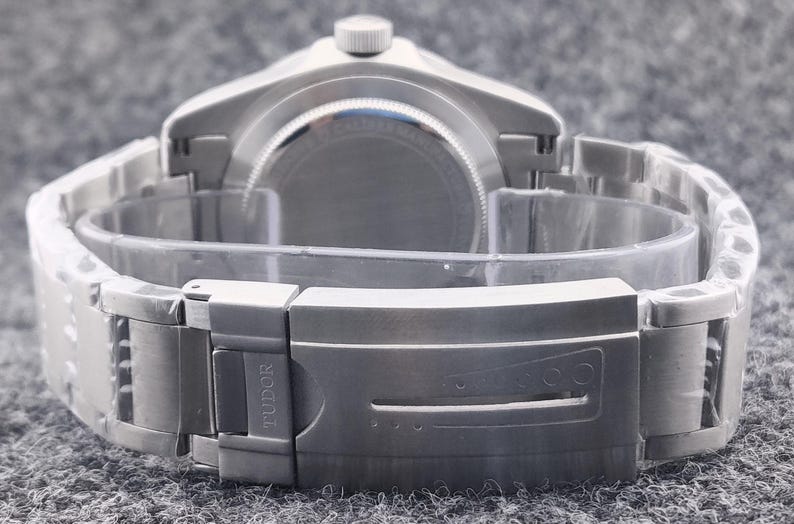 May include: A silver-toned wristwatch with a metal band and clasp. The watch is viewed from the back, showcasing the band's design and the clasp with the brand name "TUDOR" engraved. The watch is on a gray surface.