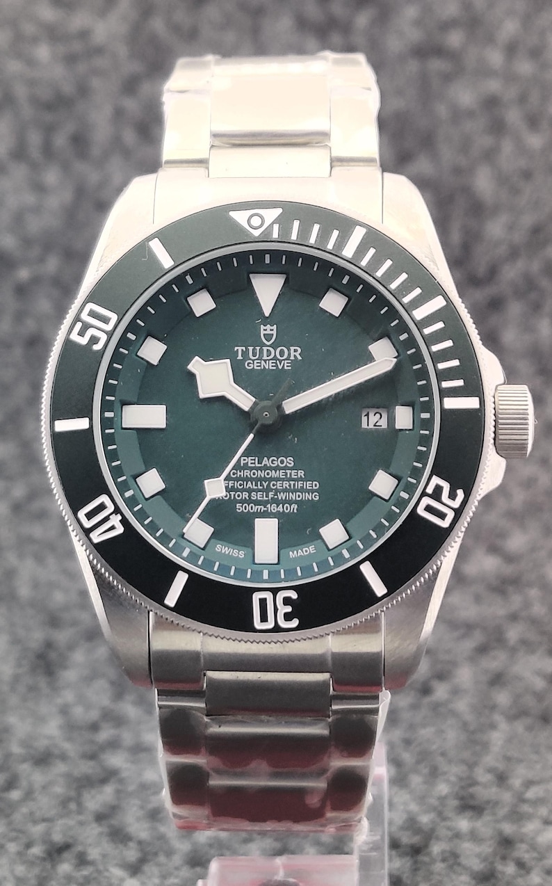 May include: A stainless steel Tudor Pelagos watch with a dark green dial and a black rotating bezel. The watch face displays the brand name, model details, and is water resistant to 500m (1640ft). The watch has a date display at the 3 o'clock position.