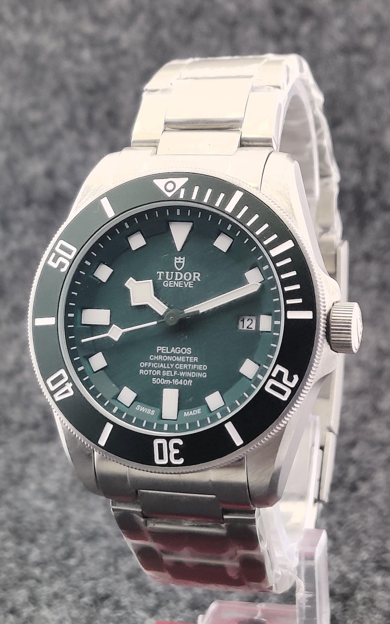 May include: A stainless steel Tudor Pelagos watch with a dark green dial and a black rotating bezel. The watch face displays the text "TUDOR GENEVE PELAGOS CHRONOMETER" and "500m-1640ft". The watch has a date display at the 3 o'clock position.