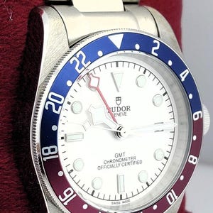 May include: A stainless steel wristwatch with a white face, a blue and burgundy bezel, and a silver band. The watch face displays the text "TUDOR GENEVE GMT CHRONOMETER OFFICIALLY CERTIFIED SWISS MADE". The watch is resting on a red surface.