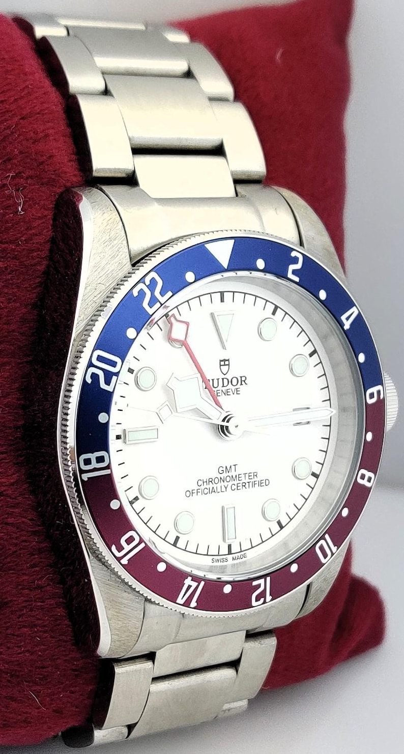 May include: A stainless steel wristwatch with a white face, a blue and burgundy bezel, and a silver band. The watch face displays the text "TUDOR GENEVE GMT CHRONOMETER OFFICIALLY CERTIFIED SWISS MADE". The watch is resting on a red surface.