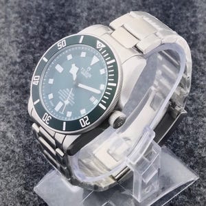 May include: A stainless steel wristwatch with a green bezel and a dark gray face. The watch has a metal band and is displayed on a clear plastic stand. The watch face displays the brand name "Tudor" and other details.