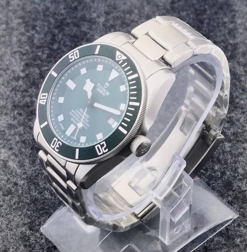 May include: A stainless steel wristwatch with a green bezel and a dark gray face. The watch has a metal band and is displayed on a clear plastic stand. The watch face displays the brand name "Tudor" and other details.