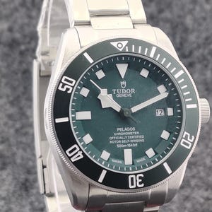 May include: A stainless steel Tudor Pelagos watch with a dark green dial and a black rotating bezel. The watch features luminous markers, a date display, and a stainless steel bracelet. The dial reads "PELAGOS CHRONOMETER" and "500m-1640ft".