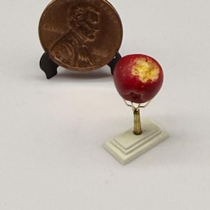 May include: A miniature red apple with a bite mark showing yellow flesh, mounted on a thin gold-colored stem and a rectangular white base. A copper-colored United States one-cent coin is displayed on a black stand behind the miniature apple.