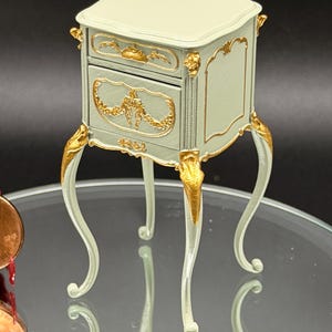 Puede incluir: Una mesita de noche en miniatura con acabado en verde salvia pálido y detalles decorativos dorados. La pieza cuenta con un cajón, una superficie superior rectangular plana y cuatro patas delgadas y curvas con detalles dorados en la parte superior.