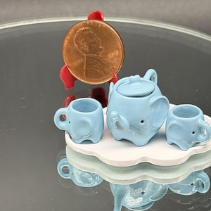 May include: A miniature light blue ceramic tea set featuring an elephant-shaped teapot and two matching elephant-shaped cups on a white scalloped tray, displayed next to a copper penny for scale to show the small size of the items.
