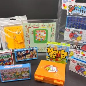 May include: A collection of miniature board games and toys including World's Smallest Connect 4, Elf-Poly, Clue, Mouse Trap, Candy Land, Faces, and an orange Artist Drawing Kit case. The items are packaged in plastic, cardboard boxes, and cases.