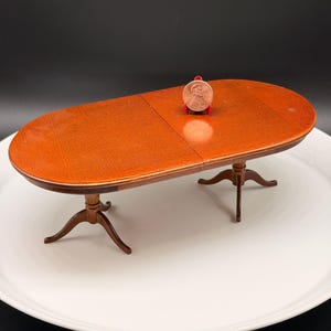 May include: A miniature oval wooden dining table with a smooth, polished brown finish. The table features two pedestal bases, each with three curved legs. A single copper-colored United States penny is placed on the tabletop for scale.