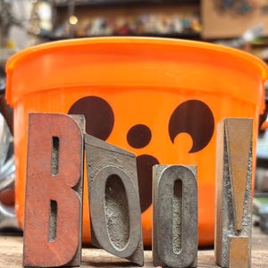 May include: Four individual vintage metal letterpress blocks arranged to spell the word BOO! in a vertical orientation. The letters are weathered with a dull grey and reddish-brown finish, standing in front of a bright orange plastic bucket.