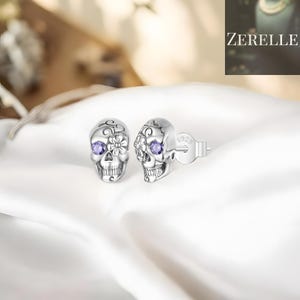 May include: A pair of silver skull stud earrings with purple gemstone eyes and floral detailing. The earrings are displayed on a white satin surface. The brand name "ZERELLE" is visible in the background.