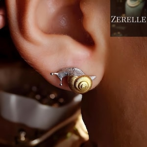 Puede incluir: Un pendiente de tuerca de caracol plateado y dorado se muestra en una oreja. El cuerpo del caracol es plateado y la concha es dorada con un diseño en espiral. La joya es única.