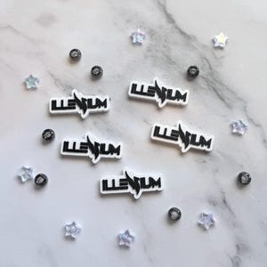 May include: Five white rectangular charms with black lettering that reads ILLENIUM, accompanied by several small iridescent star-shaped beads and dark grey circular beads scattered around the charms on a white marble-patterned surface.