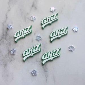 May include: Five green and white shoe charms, each featuring the text GRIZ in a stylized font, arranged alongside several small, iridescent, star-shaped decorative accents. The charms have a raised, molded design with a green base and white lettering.