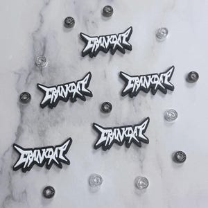 May include: Five black and white "CRANKDAT" logo patches with a bold, stylized font. The patches are scattered on a marble surface, along with small, silver and black metal embellishments. The patches have a black border and white text.