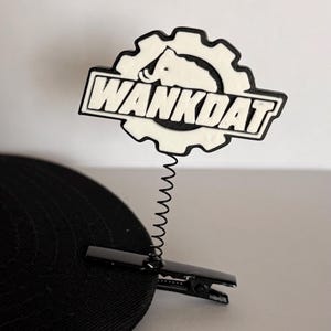 May include: A black and white hat accessory featuring a gear-shaped emblem with a mammoth silhouette and the text WANKDAT. The emblem is attached to a thin black metal spring, which is connected to a black alligator clip for secure attachment.