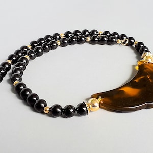 Puede incluir: Un collar de cuentas negras con un colgante grande marrón y amarillo. El collar presenta detalles dorados y un cierre. El colgante tiene una forma curva única. El collar se muestra sobre un fondo gris claro.