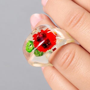Könnte beinhalten: Ein klarer, klobiger Ring aus Kunstharz mit einer eingebetteten leuchtend roten Mohnblume und kleinen grünen Blättern. Das transparente Material bildet eine glatte, abgerundete Oberfläche, die die darin eingeschlossenen botanischen Elemente zeigt.
