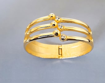 May include: A gold-coloured hinged bangle bracelet with three curved bands. Each band is accented with a small, round gold bead and a clear, sparkling stone. The bracelet is on a grey background.