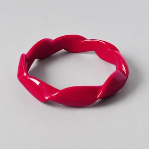 Pode incluir: Um bracelete rígido de cor rosa fúcsia brilhante com um design contínuo torcido semelhante a uma corda. O acessório possui um acabamento liso e brilhante e formato circular, construído a partir de um material sólido com uma cor uniforme e vibrante.
