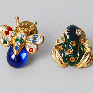 May include: Two gold-tone metal pins. One pin is shaped like a bee with clear, red, green, and blue rhinestones and a large blue teardrop-shaped stone. The other pin is shaped like a frog with green enamel, clear rhinestone eyes, and orange rhinestones.