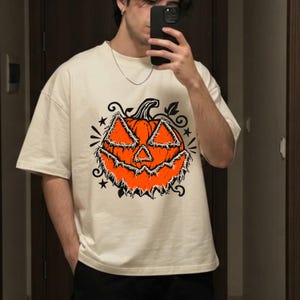 May include: A cream-colored short-sleeve t-shirt featuring a large, central graphic of a bright orange jack-o'-lantern with a distressed, frayed-edge appearance. The pumpkin design includes black outlines, swirling vine accents, and small stars.
