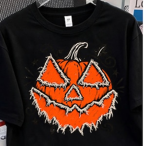 Peut inclure: Un t-shirt noir à manches courtes arborant un grand motif central représentant une citrouille jack-o'-lantern orange vif. La citrouille présente un design effiloché et usé pour ses yeux triangulaires, son nez et sa bouche dentelée.