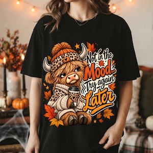 Peut inclure: Un t-shirt noir avec un motif de vache Highland portant un bonnet et un pull à imprimé léopard, tenant une tasse de café. Le design inclut des feuilles d'automne orange et jaunes, des étoiles et le texte "Not in the Mood Try again Later."
