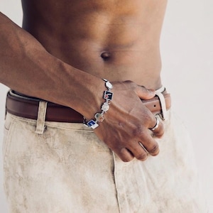 May include: Silver bracelet with various small, colorful charms. The bracelet features square, rectangular, and circular charms with black, blue, and white accents. The bracelet is worn on a wrist.