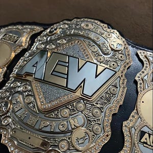 Könnte beinhalten: Nahaufnahme eines Wrestling-Meisterschaftsgürtels. Der Gürtel hat eine große, silberne und goldene Mittelplatte mit den Buchstaben "AEW". Die Worte "CHAMPION" sind auf der Platte eingraviert. Der Gürtel ist mit Strasssteinen und aufwendigen Designs verziert.