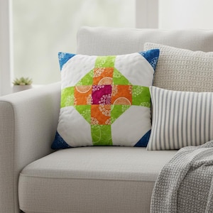 May include: A decorative square pillow with a patchwork design. The pillow features a white background with a colorful cross pattern in green, orange, and pink, with blue corner accents. The pillow is placed on a light gray sofa.