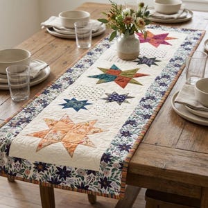 May include: A rectangular quilted table runner featuring a central cream-colored panel decorated with various multi-colored star patterns in shades of orange, blue, and green, bordered by a floral print fabric with dark blue and purple flowers.