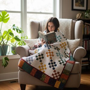 Op de afbeelding: Een persoon zit in een beige fauteuil, gehuld in een witte quilt met een kleurrijk patchwork-ontwerp. De quilt heeft vierkanten in blauw, geel en oranje. De persoon leest een boek. De sc&egrave;ne speelt zich af bij een raam met potplanten.