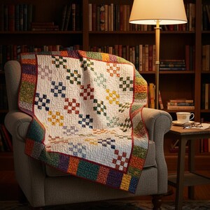 Op de afbeelding: Een handgemaakte quilt over een grijze fauteuil. De quilt heeft een witte achtergrond met kleurrijke vierkante patronen in rood, blauw, groen en geel. De randen zijn afgezet met diverse stoffen met patronen. Een lamp en een boekenkast staan op de achtergrond.