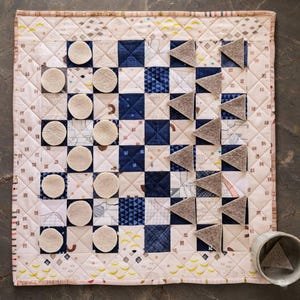 May include: A square quilted game board featuring a grid of alternating dark blue patterned squares and light cream squares with various line designs. The board includes ten circular felt pieces and ten triangular felt pieces in beige.