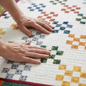 Op de afbeelding: Een handgemaakte quilt met een witte achtergrond en een herhalend patroon van kleurrijke vierkanten. De vierkanten zijn in tinten blauw, oranje, groen en geel. De quilt wordt door twee handen aangeraakt.