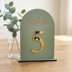 May include: An arched sage green table number sign featuring the word table in gold script and a large gold reflective number 5 attached to the front. The sign is mounted on a dark brown rectangular base and stands upright on a wooden surface.