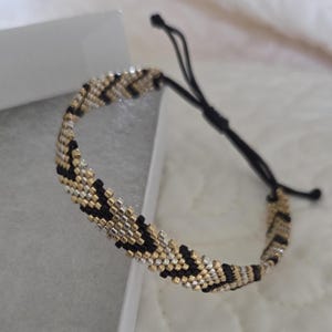 May include: Adjustable beaded bracelet featuring a geometric pattern in black, gold, and silver. The bracelet is made of small, evenly spaced beads and has a black cord closure. The bracelet is shown on a white surface.