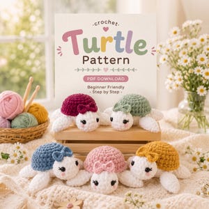 May include: A crochet turtle pattern PDF download sign featuring five small, handmade plush turtles in maroon, sage green, blue, pink, and yellow, each with a matching bow, white head, and black eyes, displayed on a wooden crate.