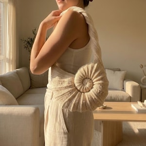 以下が含まれることがあります： クリーム色の、らせん状の貝殻のデザインの、かぎ針編みのハンドバッグ。バッグには長いストラップが付いており、肩からかけて使用します。バッグは柔らかく、質感のある糸でできており、ユニークなアクセサリーです。