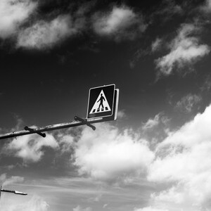 May include: Black and white photograph of a pedestrian crossing sign against a cloudy sky. The sign, mounted on a metal pole, features a white pedestrian symbol on a black background. The sky is filled with fluffy white clouds.