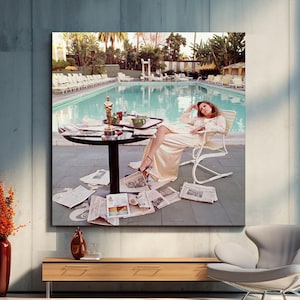 May include: A large framed print depicts a woman in a cream-colored outfit lounging by a pool. A small table with an Oscar statue and newspapers are in the foreground. The scene is set outdoors with a pool and palm trees in the background.