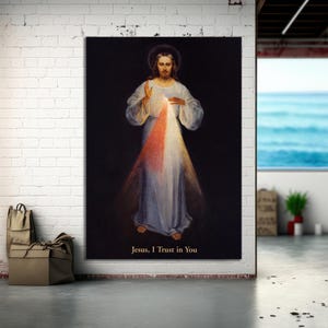 May include: A religious artwork showing Jesus with a halo, in a white robe, emitting light. The image includes the text "Jesus, I Trust in You" and features red and white rays. The background is black. The artwork is displayed on a white brick wall.