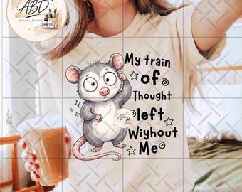 May include: A cream-coloured t-shirt featuring a cartoon illustration of a grey opossum with wide eyes. The text reads, "My train of Thought left Wiyhout Me," accompanied by small star and swirl graphics. The design is printed on the front centre.
