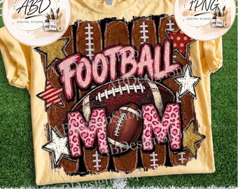 May include: A pale yellow t-shirt featuring a graphic design with the text FOOTBALL MOM. The design includes a brown football, pink leopard print lettering, and various stars in gold, red striped, and white patterns against a dark background.