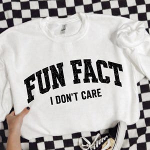 May include: White crew neck sweatshirt with the text "FUN FACT I DON'T CARE" in black, bold, distressed lettering. The sweatshirt is on a black and white checkered surface, with a black and white shoe visible.