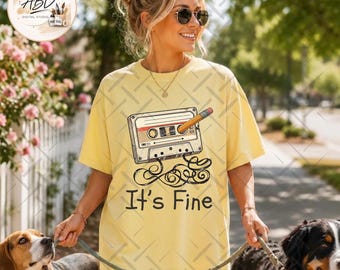 May include: A pale yellow short-sleeve t-shirt featuring a graphic of a vintage cassette tape with a yellow pencil inserted into the spool. Below the cassette, the text reads It's Fine in a dark, rounded, handwritten-style font.