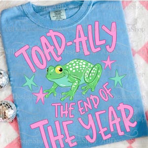 以下が含まれることがあります： 「TOAD-ALLY THE END OF THE YEAR」のピンク色の文字が入った水色のTシャツ。白い斑点と黄色の目を持つ緑色のカエルが中央にあり、ピンクと緑の星があります。