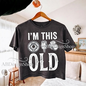 Puede incluir: Camiseta gris oscuro con el texto "I'M THIS OLD" en blanco, junto con ilustraciones de piezas de automóviles. La camiseta presenta un componente de freno redondo, un distribuidor y un componente de estrangulador. La camiseta está colgada en una percha de madera.